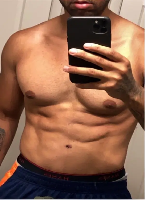 A person takes a mirror selfie, showing their muscular upper body and abs, while wearing dark shorts.